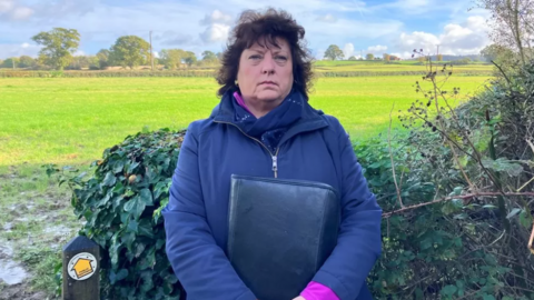 Jo Kendall, wearing a blue jacket and holding a bag close to her stomach, standing in front of a field