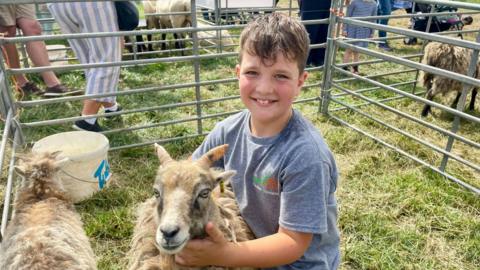 A boy in a grey t-shirt kneels on the grass hugging a sheep. The boy has short brown hair and his T-shirt has a logo which reads NANTWICH SHOW. The sheep has short horns and caramel coloured wool. The pair sit in a gated pen with a bucket and another sheep. Spectators can be seen walking in the background