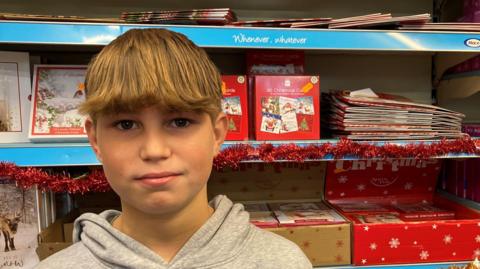 Ethan standing in front of a Christmas shelf in Kinnerley Shop. There are Christmas cards and tinsel behind him. 