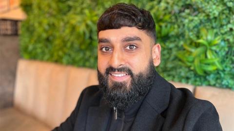 Close up of Usman Hussain in the cafe, he has short dark hair and a beard and is wearing a black smart coat