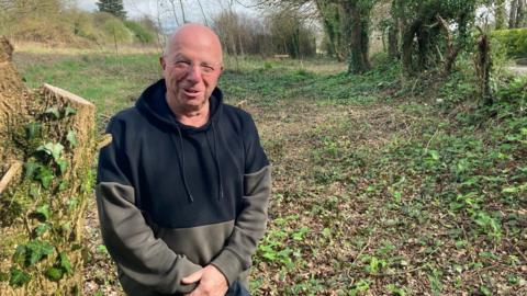 A man, wearing a black and green hoodie, with a bald head and glasses, stands in front of a clearing lined with trees near Curry Rivel on the Somerset Levels.