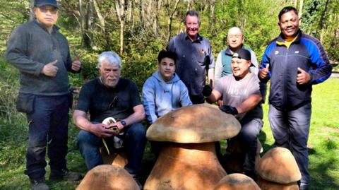 A group of men stand in a woodland area alongside large wooden toadstools.