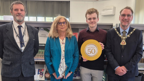 Four people dressed smartly are standing together in front of book shelves in the library.  One man holds an orange circle which reads - Yaxley Library is 50.