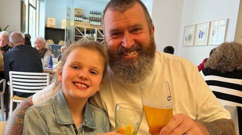A girl wearing a denim jacket is sitting next to a man wearing a white t-shirt. He has a beard and tattoos. Both are holding a drink in a glass. They are in what appears to be a restaurant with other people sitting at tables behind them.