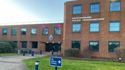 The front of the building of Bedfordshire Police headquarters. It is a red brick building with grass and bushes outside. In the foreground there is a sign for visitors parking.