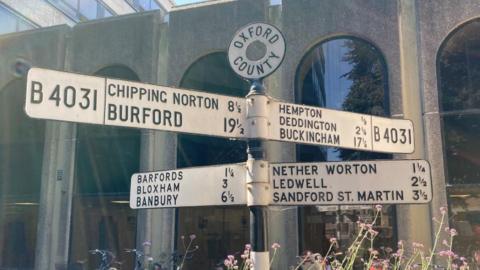 A road sign with lots of different place names and how far away they are. It's in front of a building with large arched windows.