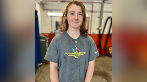 Mobile mechanic Jess Barker smiling for a picture. She is wearing a grey T-shirt with an Indianapolis Motor Speedway logo on. There are several machines behind her.
