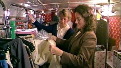  Two women looking at dresses in a clothes shop which is on a double-decker bus.