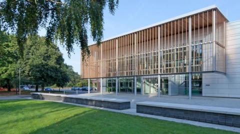An exterior view of Havant Borough Council's main building, the Public Service Plaza. It is a modern-looking building, made with large white brick and has an artistic panelling aorund the exterior made of pale wooden slats. The building has a car park to the left and is surrounded by a patch of grass and some trees.
