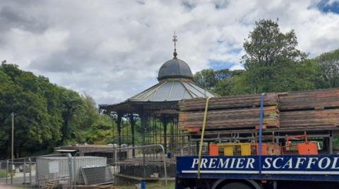 Scaffolding being erected at the fenced-off bandstand. A lorry with building materials is parked in front of it. The structure has ornamented railings and a grey roof with a cupola on top of it. It is surrounded by trees.