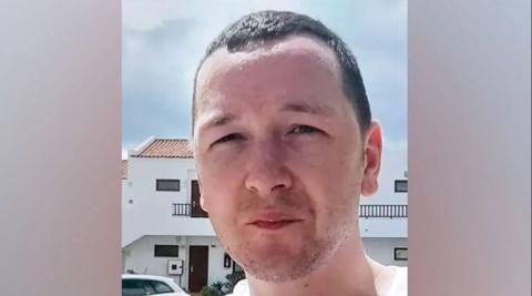 A man with short dark hair and a T-shirt looks at the camera, with a white apartment building in the background