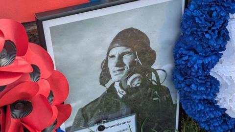 A close up framed picture of Czech pilot Alois Vasatko placed on the side a memorial made in his memory. There is a poppy wreaths and tributes next to it.