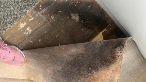A carpet lifted to show its underside, which is black with damp. A pink trainer is holding the carpet in place.