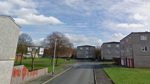Street view of a residential estate with several grey three-storey apartment blocks on both sides of a quiet road. A grassy area and footpaths line the street, with a few leafless trees and a lamppost visible. The sky is partly cloudy with patches of blue, and no people or vehicles are present. Faint graffiti can be seen on a low wall in the left foreground.