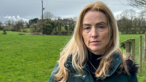 Colette Casey has long blonde hair and is wearing a dark green coat. She is standing in a field, behind her a hedgerow and trees can be seen as well as a telegraph pole.