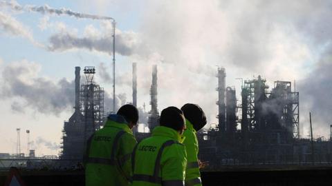Three people in yellow high visibility jackets and black woollen hats walking in front of the smoking chimneys of the Lindsey Oil Refinery