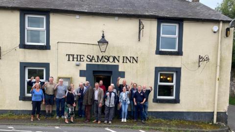 A group of 17 people raising a glass outside a cream coloured building. Words spell out 'The Samson Inn' above the front door. 