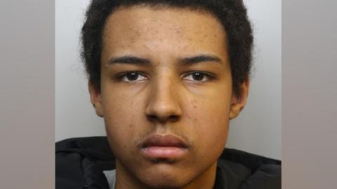 A mugshot of Romeo Dumbi. He has short black hair and some stubble on his upper lip. He is pictured looking directly at the camera with a blank expression.