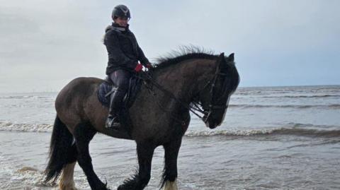 Harriet Martin and Guinness walking along the coastline in the shallow water. Guinness is looking down and ahead to the right and Martin is looking into the camera and smiling.