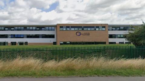 A modern, rectangular school building with three visible floors. The structure has a mix of white and light brown brick sections. The building features large horizontal rows of windows across all floors.
In the centre-right section of the building, there is a prominent sign with the logo and name “Outwood Academy Adwick” displayed on the brown brick portion.
The building is set back from the road behind a green metal fence, and in front of the fence, there is a strip of tall, dry grass. The foreground shows a paved road running parallel to the fence.