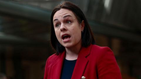 Kate Forbes, who has short dark hair, speaks with her mouth wide open. She is wearing a red jacket over a blue top. 
