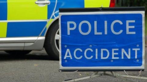 A close up of a blue rectangular sign, resting on tarmac, saying police accident in white letters. Behind it is a close-up of the rear view of a police car, marked with blue and yellow rectangles. 