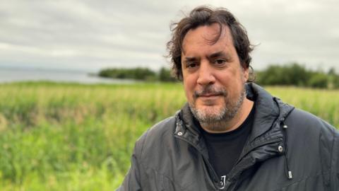 Dr. Eduardo Salazar-Ortuño. He has dark hair and grey beard, wearing a black raincoat and black t-shirt. He is standing on the shores of Lough Neagh.