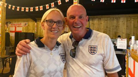 A woman and man in England shirts are smiling while standing inside The Big Barn.