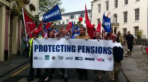 A white banner with the words protecting pensions being held by a number of people at the front of a column of protestors holding banners. 