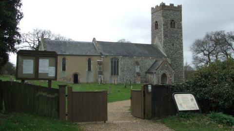 Church with a square tower in a church yard. There are gravestones in front of the building which is also grassed and a gravel path leads to the entrance. A low wooden fence skirts the area.