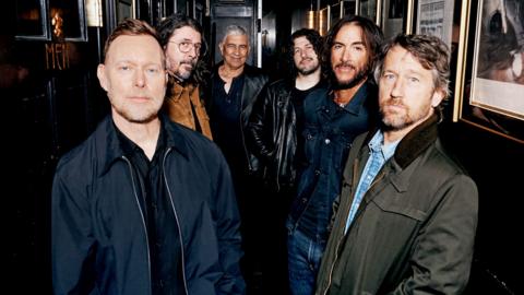 Six members of the Foo Fighters band stand side by side looking directly at the camera, hands in pockets.