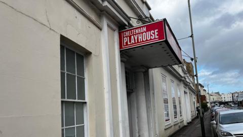 An exterior view of the Cheltenham playhouse showing a red sign with the theatre's name on it. The building is painted white.