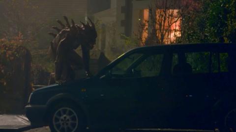 Shadowy monster with spikes sticking out of back crawls up a car in front of a house during the night.