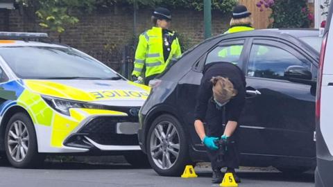 A forensics officer taking photos of a crime scene. There is a black car and a police car behind the officer, as well as two police officers.