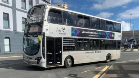 A silver double decker bus driving through Douglas. The digital sign at the front has orange writing and the Bus Vannin logo with the name of the company and a triskelion can also be seen on its side.