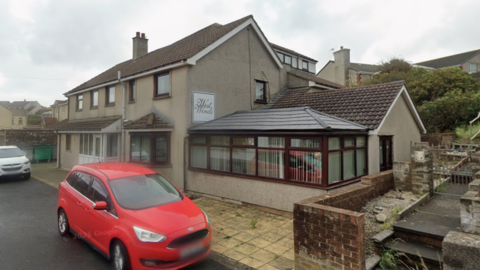 Westwinds Residential Care in Workington is a grey building with a conservatory attached to it. It has a small patio and two cars parked in front of it. It is a wet grey day. 