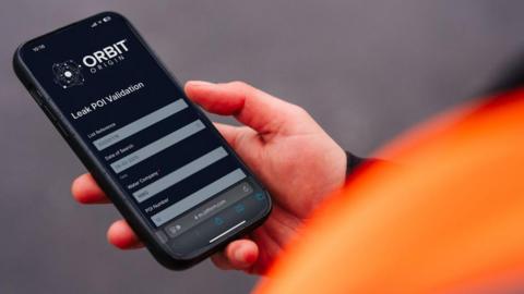 A person's zoomed-in hand holding a black smartphone which displays an app. The apps title is Orbit Origin. There is a blurred orange jacket to the left of the picture along with a blurred grey background.