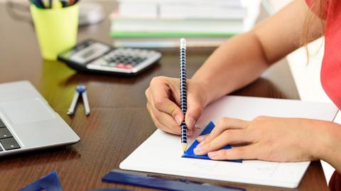 GCSE maths image: a student sits at a table drawing a line in a notebook using a triangular ruler.