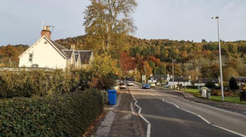 A leafy Highland village scene, with traditional, white-walled properties, hedges and trees lining a road. In the distance is a low, tree-covered hill.