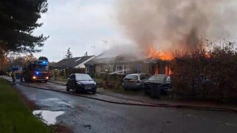 A bungalow on fire. The property is well alight with flames and smoke. Fire crews wearing protective clothing and hard hats use hoses to spray water on to the building. A fire engine can also be seen.