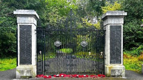 A set of memorial gates with a marble pillar either side. At the base of each post is a gap where there was once a plaque