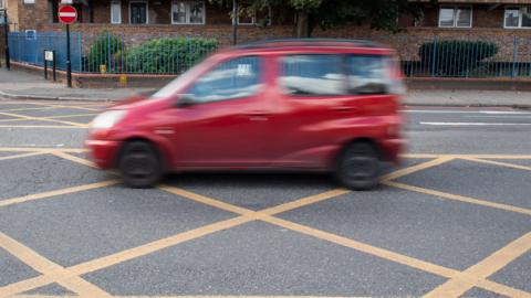Small red car blurred out passing through a yellow box junction on a main road.