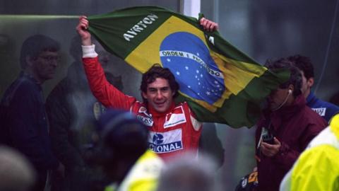 Ayrton Senna celebrates winning the European Grand Prix at Donnington in 1993, holding a Brazilian flag behind his head.