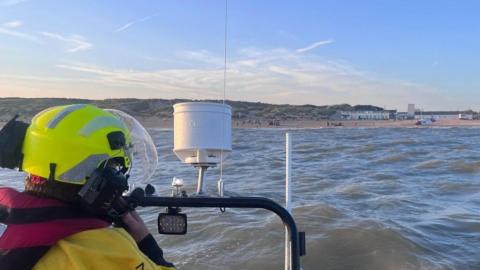 A picture taken from a lifeboat looking back to land, with a man on it making a phone call.