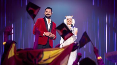 Rylan and his co-host of the Interstellar Song Contest in Doctor Who stand on stage as people wave flags in the air
