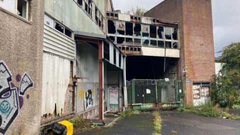 Derelict former factory site - shows bins overturned, graffiti.