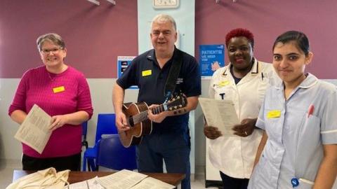 A man with grey hair is holding a guitar. Around him are three women who work at the hospital, one dressed in light bur scrubs, the other in white uniform, and the other in a pink jumper.