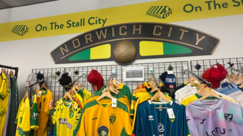 Different coloured football shirts and baseball caps are hung up on hangers in rows on a wall. There is a sign above them which says NORWICH CITY in gold writing with a green and yellow logo. The shop's logo On The Stall City is on a banner round the top of the shop in yellow and green writing.