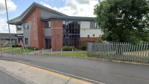 A GP surgery in Bradford, with the word "Farrow" written vertically down the red brick building. In the foreground there is a wide pavement, several bollards and a large tree behind a metal fence. 