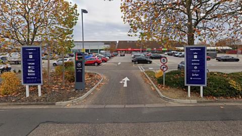 The entrance to Ortongate Shopping Centre, showing a car park and shops in the background. Leaves are falling off the trees.